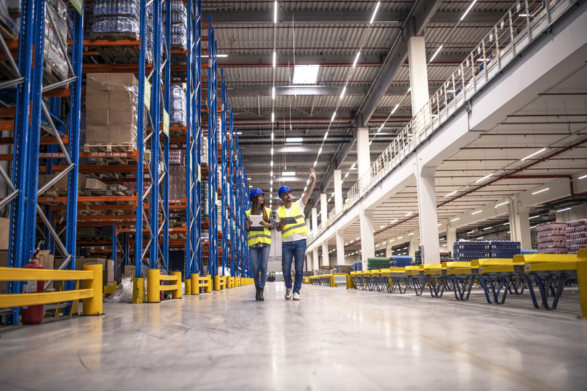 Distribution warehouse interior with workers wearing hardhats and reflective jackets walking in storage area.